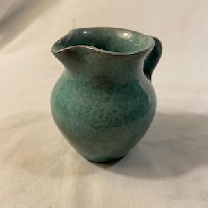 May include: A small, teal-colored ceramic pitcher with a rounded body and a small handle. The pitcher has a slightly irregular rim and a speckled glaze, giving it a handmade appearance. The overall design is simple and functional.