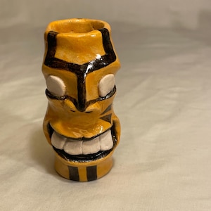 May include: A ceramic tiki mug with a smiling face. The mug is primarily yellow with black and white accents. The design includes a wide grin with white teeth and black stripes around the top and bottom.