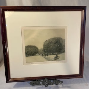 May include: A framed print of a park scene with a dark brown wooden frame. The artwork depicts a park with trees, a path, and figures. The print is matted with a white border. The frame is supported by a decorative metal stand.