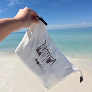 May include: A white canvas bag with a black zipper and a black leather pull tab. The bag features a brown illustration of a bikini hanging on a clothesline, with the name "abigail" printed below. The background is a beach with blue sky and water.