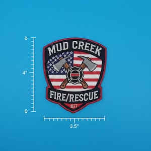 Op de afbeelding: Geborduurde patch met de tekst "MUD CREEK FIRE/RESCUE 1977". De patch heeft een schildvorm met gekruiste bijlen en een ontwerp van de Amerikaanse vlag. De patch is ongeveer 10 cm hoog en 9 cm breed.
