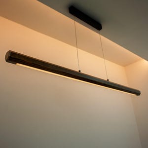 Op de afbeelding: Een lineaire hanglamp met een lange, zeshoekige behuizing van donker hout, opgehangen aan twee dunne metalen kabels vanaf een zwarte rechthoekige plafondbevestiging, met een geïntegreerde LED-strip die warm licht naar beneden uitstraalt.