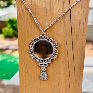 May include: A silver-toned pendant necklace featuring a circular mirror framed by an ornate, scrollwork metal design that resembles a handheld mirror. The pendant hangs from a matching silver-toned chain, showcasing intricate detailing on the handle.