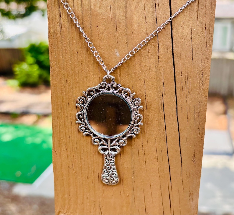 May include: A silver-toned pendant necklace featuring a circular mirror framed by an ornate, scrollwork metal design that resembles a handheld mirror. The pendant hangs from a matching silver-toned chain, showcasing intricate detailing on the handle.