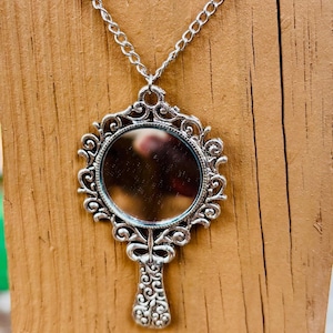 May include: A silver-toned pendant necklace featuring a circular mirror framed by an ornate, scrollwork metal design. The pendant is shaped like a handheld mirror with a decorative handle and is suspended from a matching silver-toned metal chain.