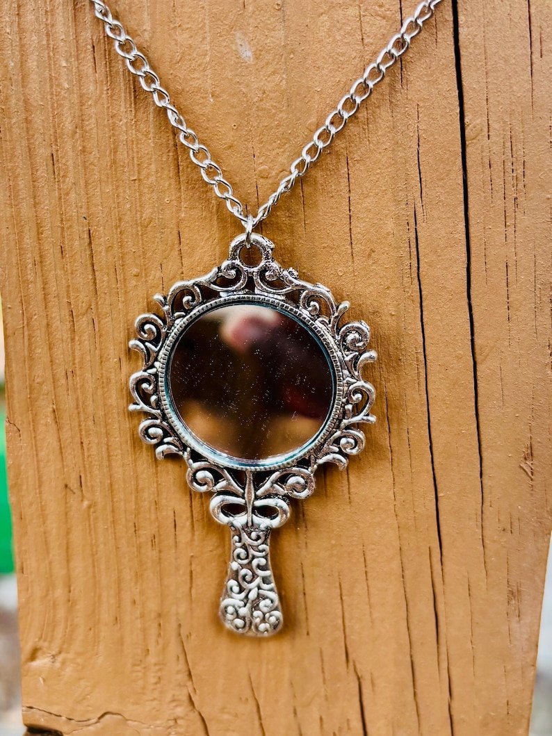May include: A silver-toned pendant necklace featuring a circular mirror framed by an ornate, scrollwork metal design. The pendant is shaped like a handheld mirror with a decorative handle and is suspended from a matching silver-toned metal chain.