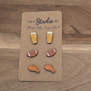 May include: Three pairs of small wooden stud earrings attached to a tan display card labeled DBM Studio '87 Perfectly Imperfect. The designs include two beer glasses with foam, two brown American footballs with white laces, and two chicken drumsticks.
