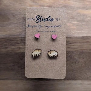 May include: Two pairs of stud earrings on a brown display card labeled DBM Studio '87 Perfectly Imperfect Sterling Silver Plated. One pair features small pink wooden hearts, and the other pair features small wooden bison silhouettes with dark edges.