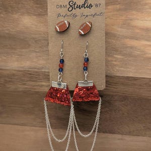 May include: A set of earrings on a display card labeled DBM Studio '87 Perfectly Imperfect. The set includes two small brown football-shaped stud earrings and two dangling earrings featuring red glitter trapezoids, blue and red beads, and silver chains.