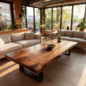 May include: A large, rectangular wooden coffee table with a natural edge and a dark, glossy finish. The table is supported by a black metal base. A small, dark bowl sits on the table's surface. The room features neutral-toned sofas and large windows.