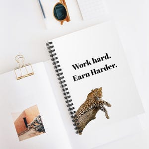 Op de afbeelding: Wit spiraalgebonden notitieboek met de tekst "Work hard. Earn Harder." en een luipaardillustratie. Een goudkleurige paperclip en zonnebril zijn ook zichtbaar.