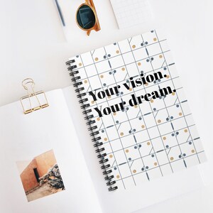 Op de afbeelding: Een spiraalgebonden notitieboek met een geometrisch patroon van blauwe en gouden stippen en lijnen op een witte achtergrond. De tekst "Your vision. Your dream." staat op de kaft. Een gouden paperclip en zonnebril zijn ook zichtbaar.