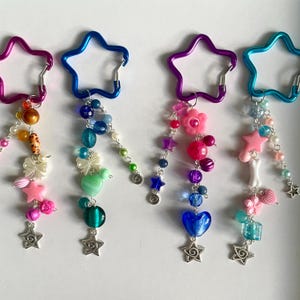 May include: Four keyrings with star-shaped carabiner clips. Each keyring has a unique design of beads, charms, and star-shaped pendants. The colour schemes include pink, blue, purple, and teal, with various bead shapes and sizes.