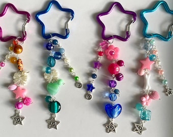 May include: Four colorful keychains with star-shaped carabiner clips. Each keychain features a unique design of beads, charms, and star-shaped pendants. The color schemes include pink, blue, purple, and teal, with various bead shapes and sizes.