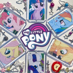 May include: A selection of My Little Pony character keyrings. Each keyring features a different pony, surrounded by decorative beads, charms, and star-shaped accents. The My Little Pony logo is in the centre, with a red apple charm.