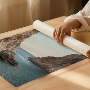 May include: A landscape print featuring a coastal scene with turquoise water, rocky cliffs, and a boat. The print is being unrolled on a wooden surface. The print's colors include shades of blue, brown, and green.
