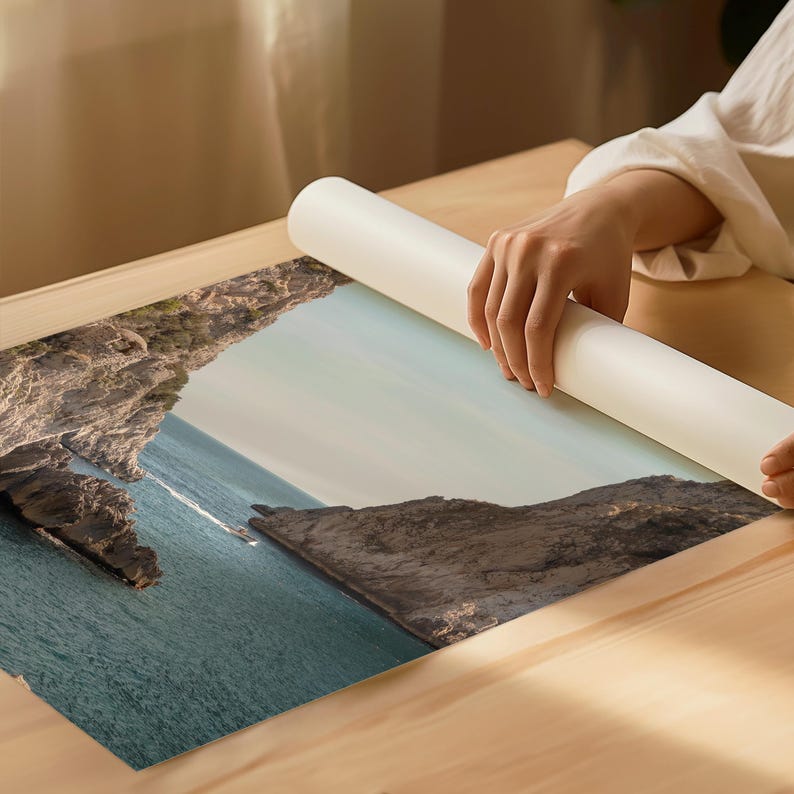 May include: A landscape print featuring a coastal scene with turquoise water, rocky cliffs, and a boat. The print is being unrolled on a wooden surface. The print's colors include shades of blue, brown, and green.