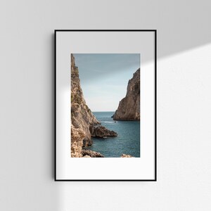 May include: A framed photograph of a coastal scene. Two large, light-colored rock formations frame a view of the blue sea and sky. A small boat is visible in the distance, adding a sense of scale to the natural landscape.