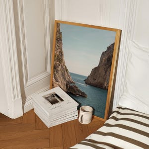 May include: A framed landscape print featuring a coastal scene with towering cliffs and turquoise water. A stack of white books with the word "JOURNAL" and a small ceramic mug are in front of the print. The frame is a light wood color.