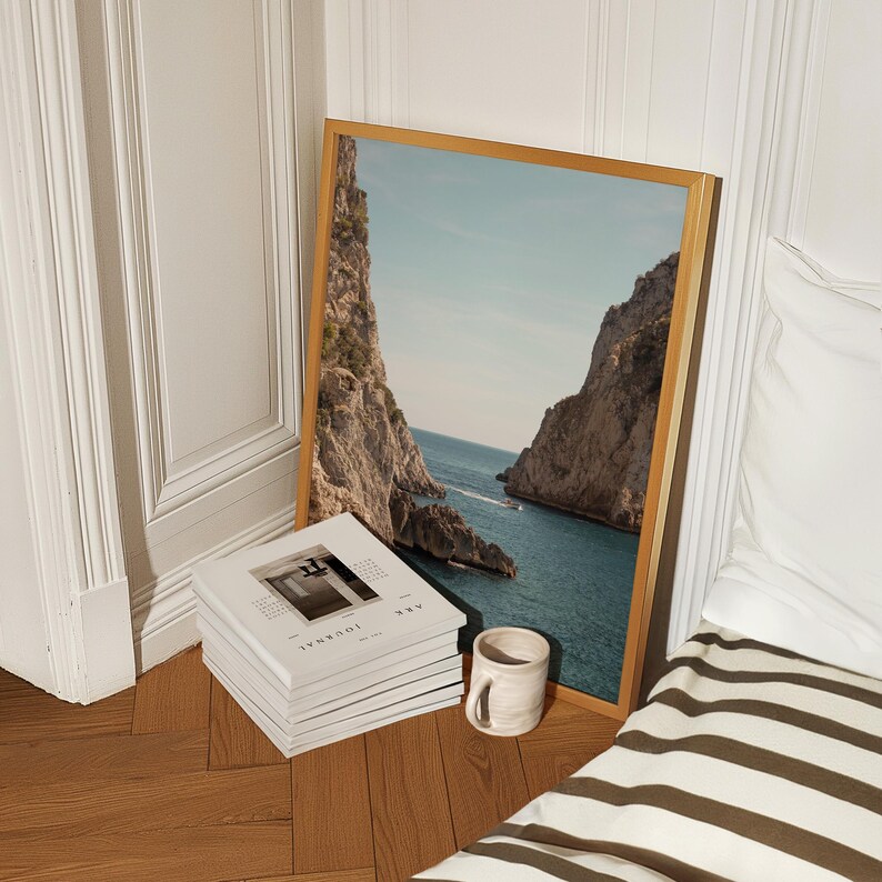 May include: A framed landscape print featuring a coastal scene with towering cliffs and turquoise water. A stack of white books with the word "JOURNAL" and a small ceramic mug are in front of the print. The frame is a light wood color.