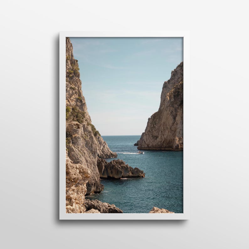 May include: A framed print featuring a scenic coastal view. The image captures a serene seascape with towering rock formations on either side, framing a view of the ocean and a boat. The water is a deep blue, and the sky is clear.