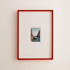 May include: A framed photograph featuring a scenic coastal view. The photograph is set within a white mat and a vibrant red frame. The image captures a view of the sea between two rocky cliffs under a blue sky.