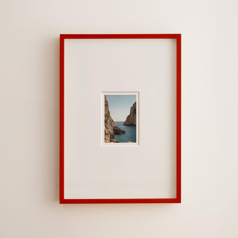 May include: A framed photograph featuring a scenic coastal view. The photograph is set within a white mat and a vibrant red frame. The image captures a view of the sea between two rocky cliffs under a blue sky.