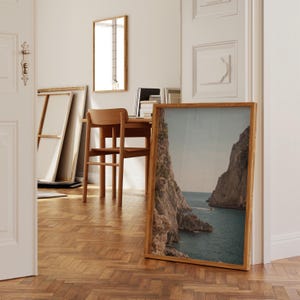 May include: A framed landscape print of a coastal scene with rocky cliffs and turquoise water. The print has a natural wood frame and is leaning against a wall. The room also features a wooden desk, chair, and mirror.