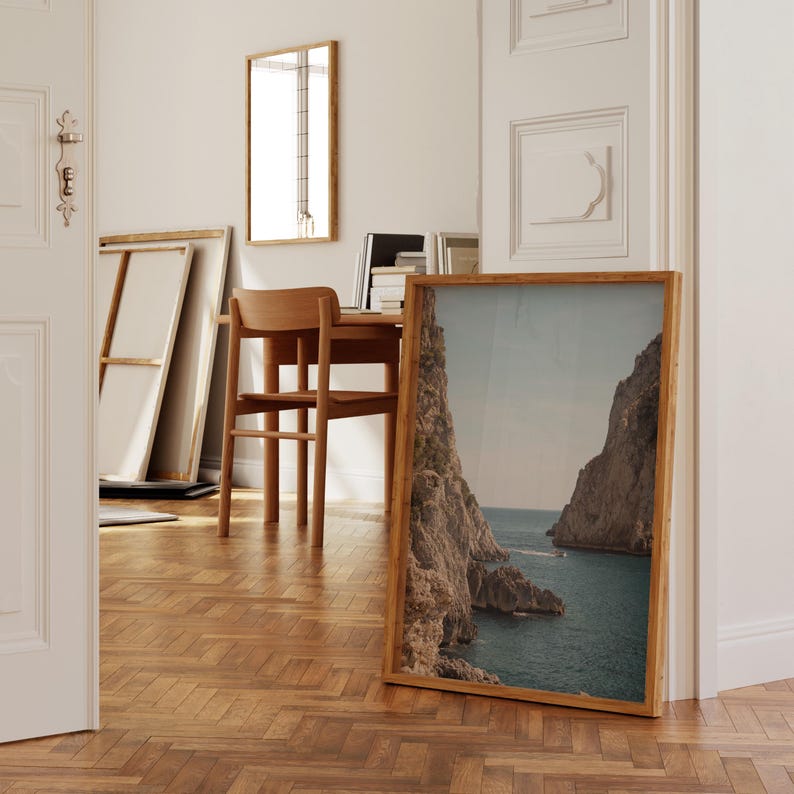 May include: A framed landscape print of a coastal scene with rocky cliffs and turquoise water. The print has a natural wood frame and is leaning against a wall. The room also features a wooden desk, chair, and mirror.