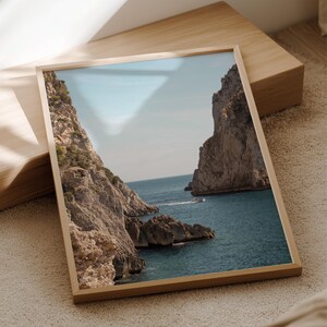 May include: A framed print of a coastal landscape featuring towering rock formations on either side of a turquoise sea. A small boat is visible in the distance. The print is in a light wood frame, resting on a wooden block.