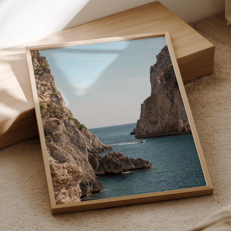 May include: A framed print of a coastal landscape featuring towering rock formations on either side of a turquoise sea. A small boat is visible in the distance. The print is in a light wood frame, resting on a wooden block.