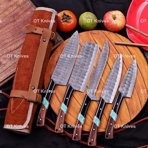 May include: A set of five kitchen knives with hammered metal blades and handles featuring dark wood and turquoise-colored inlays, arranged on a round wooden board next to a brown leather knife roll, a persimmon, a lime, and a red apple.