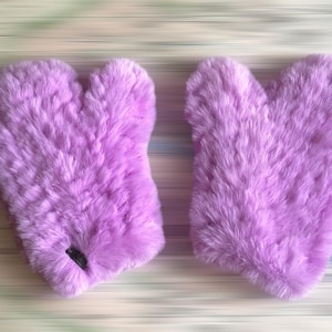 May include: Two fluffy, lavender-colored mittens. The mittens are made of a soft, textured material, likely fur. The mittens are heart-shaped, with a thumb and four-finger section. The background is a blurred, light-colored surface.