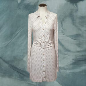 May include: A long-sleeved, button-down dress on a mannequin. The dress is a light beige color with a subtle floral pattern. It features a collared neckline and ruching detail at the waist. The dress is knee-length.