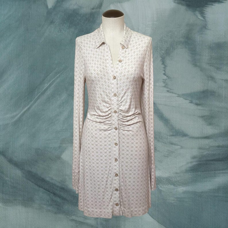 May include: A long-sleeved, button-down dress on a mannequin. The dress is a light beige color with a subtle floral pattern. It features a collared neckline and ruching detail at the waist. The dress is knee-length.