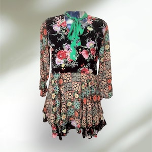 May include: A long-sleeved dress with a floral print on a black background. The dress has a green tie at the neckline and a tiered skirt with a patchwork design. The sleeves have a ruffled cuff.