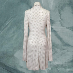 May include: A long-sleeved dress with a small, repeating geometric pattern in white and light brown. The dress is displayed on a mannequin against a teal background. The dress has a collared neckline and a flared skirt.