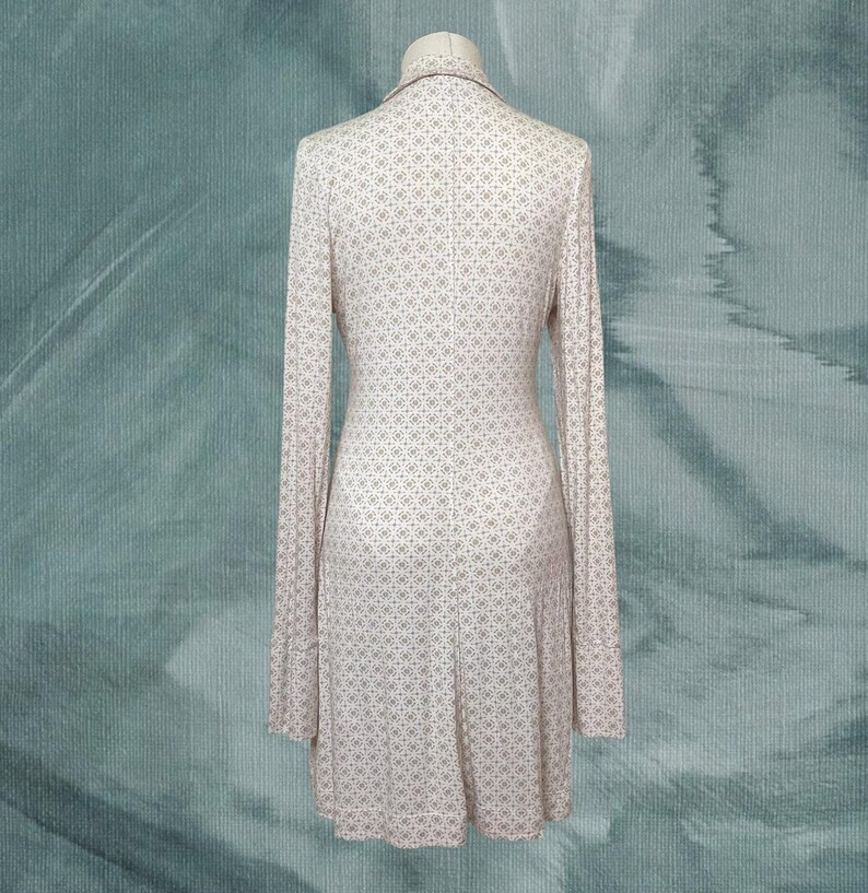 May include: A long-sleeved dress with a small, repeating geometric pattern in white and light brown. The dress is displayed on a mannequin against a teal background. The dress has a collared neckline and a flared skirt.