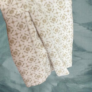 May include: Close-up of a cream-colored fabric sleeve with a repeating pattern of tan floral and geometric shapes. The sleeve is folded, revealing the interior fabric. The background is a textured teal color.