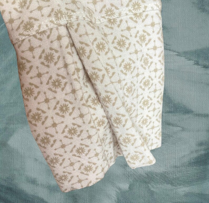May include: Close-up of a cream-colored fabric sleeve with a repeating pattern of tan floral and geometric shapes. The sleeve is folded, revealing the interior fabric. The background is a textured teal color.
