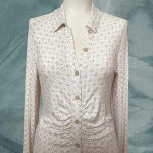 May include: A long-sleeved button-down shirt with a collared neckline. The shirt features a repeating geometric pattern in beige and white. The buttons are a light brown color. The fabric is gathered at the waist.