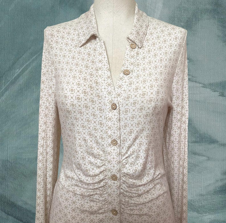 May include: A long-sleeved button-down shirt with a collared neckline. The shirt features a repeating geometric pattern in beige and white. The buttons are a light brown color. The fabric is gathered at the waist.
