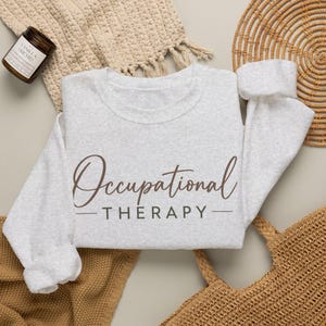 Może przedstawiać: Jasnoszara bluza z okrągłym dekoltem z nadrukiem Occupational Therapy w kolorze ciemnobrązowym. Słowo Occupational jest napisane kursywą, natomiast słowo THERAPY jest zapisane pogrubioną, wielką czcionką bezszeryfową.
