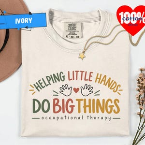 May include: An ivory t-shirt with a graphic print that reads HELPING LITTLE HANDS DO BIG THINGS occupational therapy in green, red, and yellow text, featuring two hand outlines and a small heart icon centered above the text.