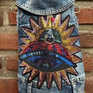 May include: A denim dog vest with a large, colorful sequined patch featuring an eye design within a sunburst. The vest has a collar and is hanging on a gold hanger against a brick wall.