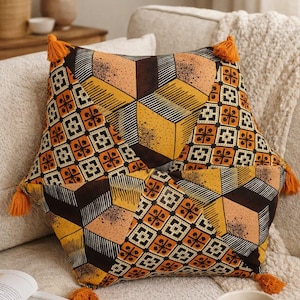 May include: Geometric patterned decorative pillow with orange tassels. The pillow features a repeating pattern of brown, orange, and yellow geometric shapes. The pillow is on a cream-colored sofa.