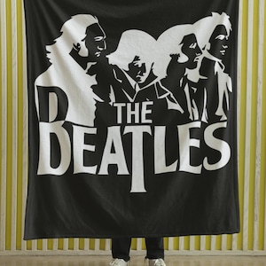 May include: Black blanket featuring a white graphic of The Beatles band members. The band's name is displayed in large white letters. The blanket is held up against a yellow and white striped background.