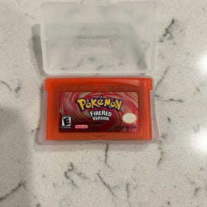 May include: An orange Game Boy Advance cartridge for the game "Pokemon FireRed Version" is displayed inside a clear plastic case. The game cartridge has the Pokemon logo and the Nintendo logo.
