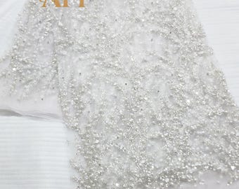 May include: White beaded fabric with a delicate, intricate design. The fabric is covered in small, shimmering beads, creating a textured surface. The letters "AFI" are visible in the upper left corner. This fabric is suitable for bridal wear or special occasion garments.