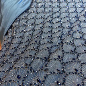 Könnte beinhalten: Nahaufnahme eines hellblauen Stoffes mit aufwendiger Perlenarbeit. Das Design zeigt ein sich wiederholendes Muster aus fächerartigen Formen, die mit kleinen, klaren Perlen umrandet und mit größeren, dunkelblauen Edelsteinen durchsetzt sind. Ein Abschnitt aus hellblauem Satin ist sichtbar.